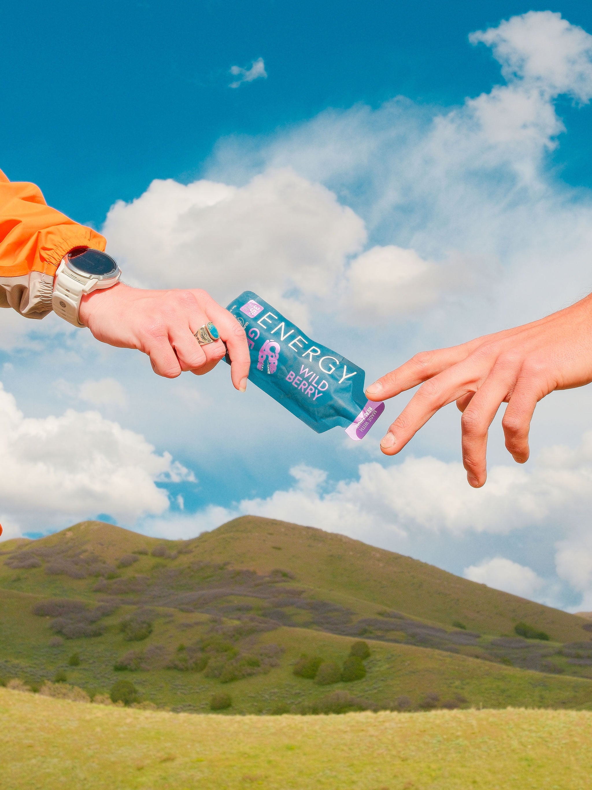 Two hands reaching towards each other with a blue energy gel package against a scenic background.